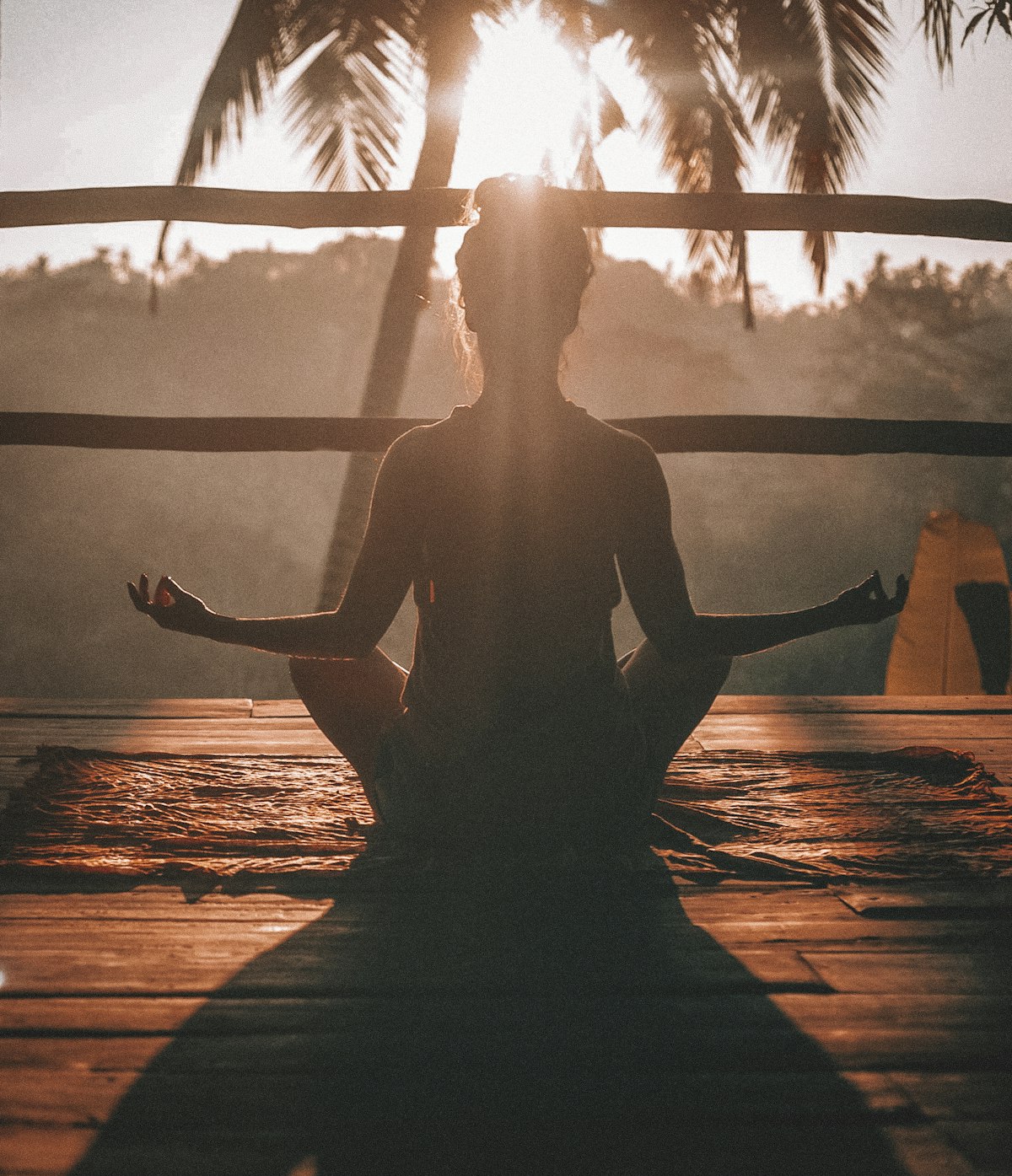 Person meditating on a wooden deck at sunrise, framed by palm trees